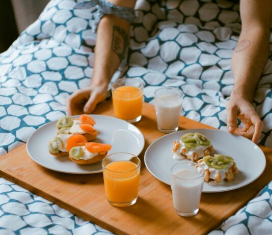 여행중에도 건강관리 하기 person serving pastries on white ceramic plates with fruit juice glasses on wooden tray on top of bed